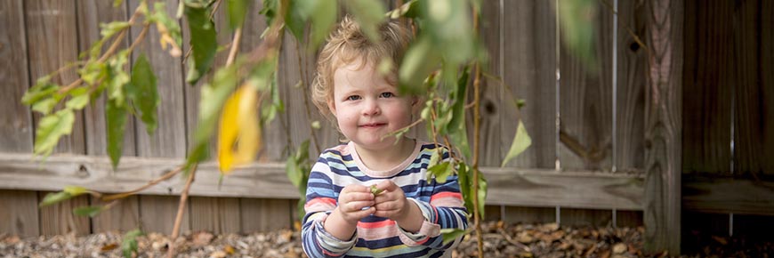A girl stands behind the leaves of a tree in the outdoor play area at the Child Development Center.