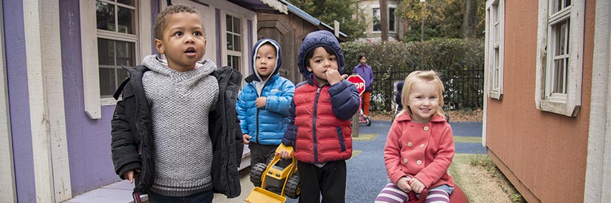 Four children outside on the playground area at the Child Development Center. Four boys stand to the left, one of whom is holding a toy truck. A girl sits on a red ball on the right. A teacher is in the background.