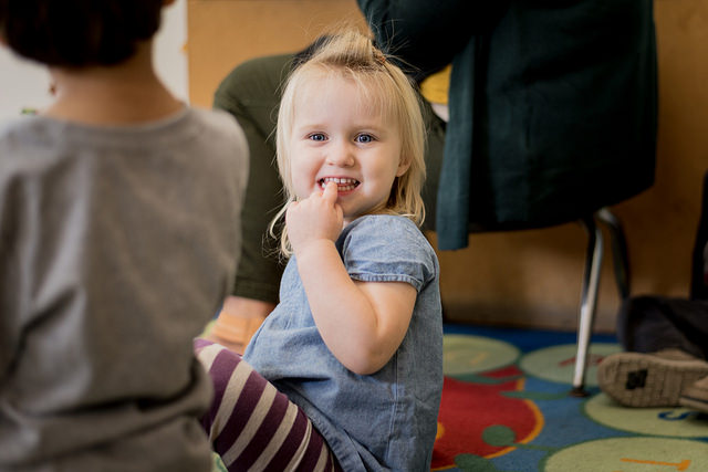 A student sits smiling with her finger in her mouth in Classroom 102 at the Child Development Center.