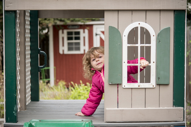 A student in a red coat at the Child Development Center looks out the door of a playhouse in the exterior playground.