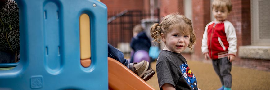 A girl sits on a slide. Another waits behind her to go down the slide. A boy in the background watches.